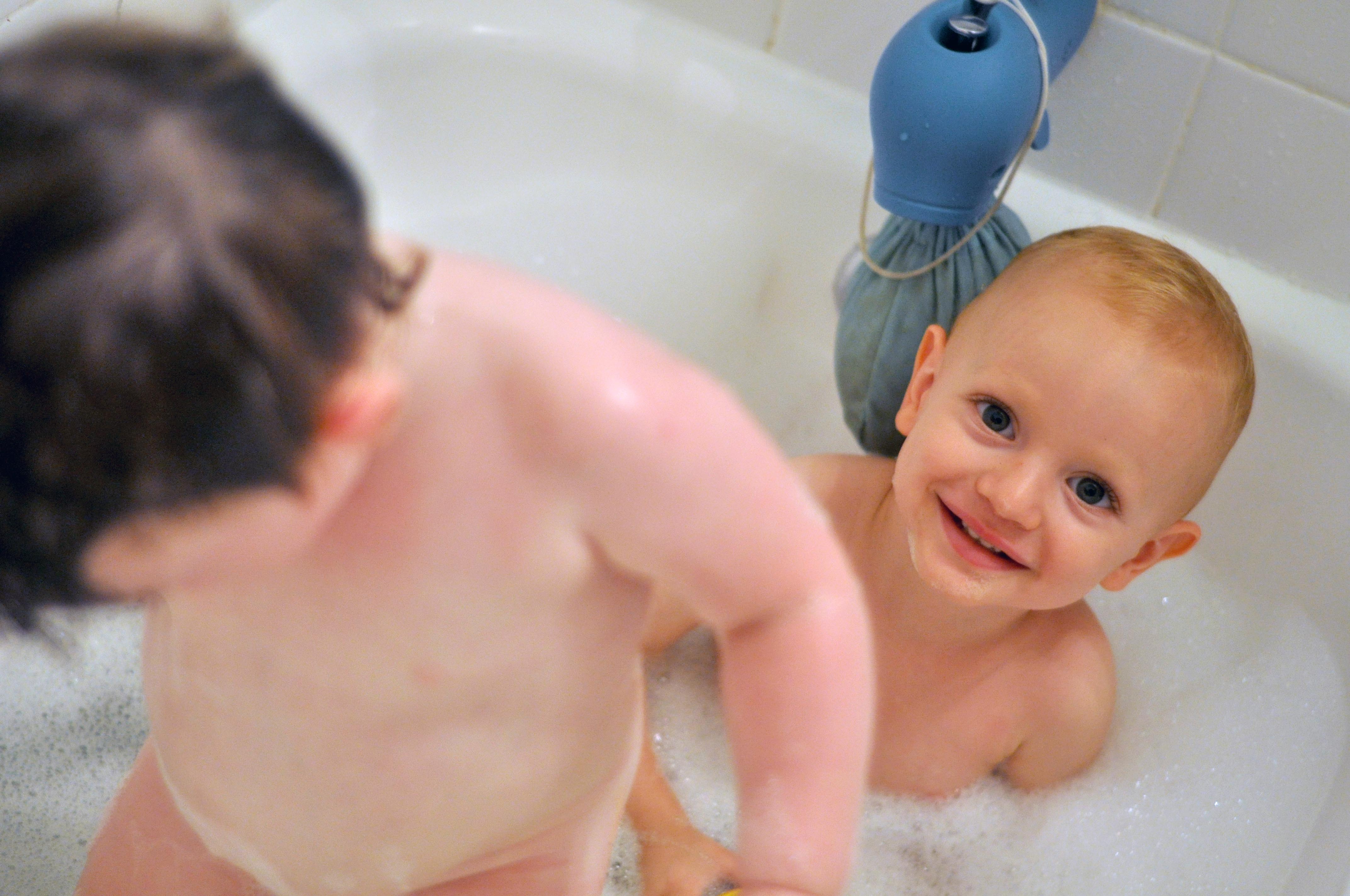 Two young children sitting in a bathtub with bubbles, one smiling at the camera.