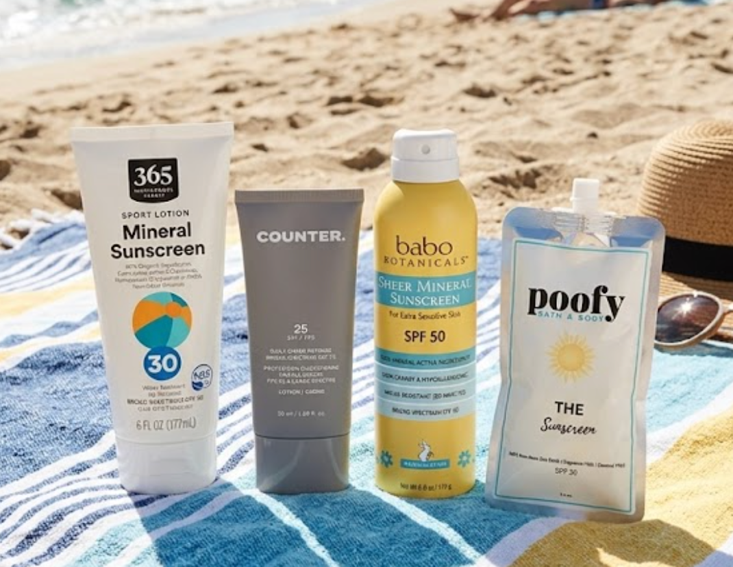 Four sunscreen products, including 365 Mineral Sunscreen, a Counter tube, Babo Botanicals spray, and a Poofy pouch, sitting together on a blue and yellow striped beach towel with the ocean in the background.