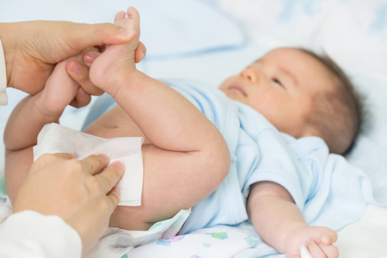Parent using baby wipes during diaper change with smiling infant on soft bedding.