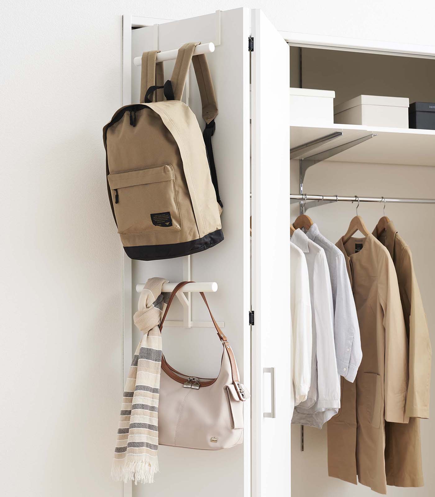 Backpack and handbag hanging on a white wall-mounted rack with clothes on hangers inside a closet.