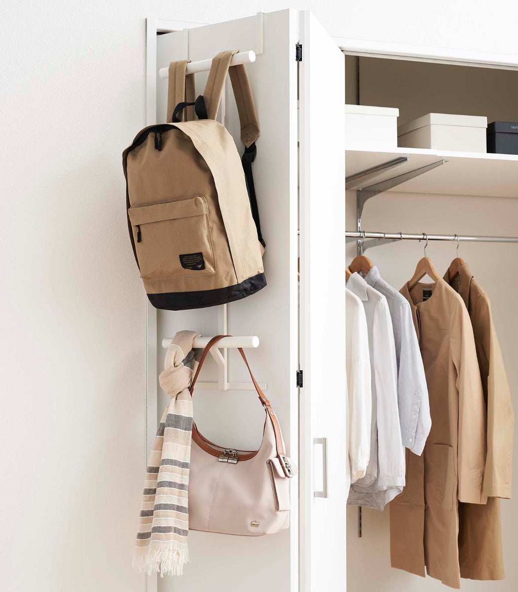 Backpack and handbag hanging on a white wall-mounted rack with clothes on hangers inside a closet.