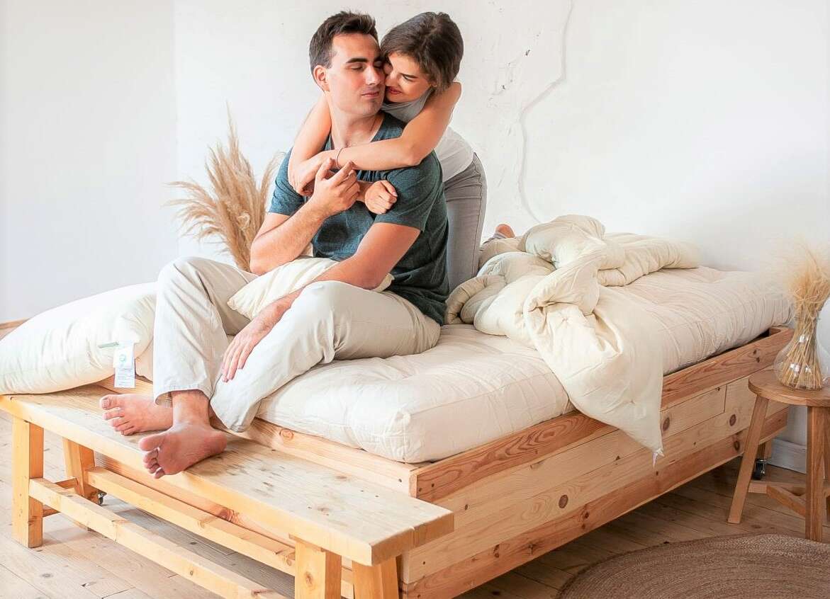 Couple embracing on a wooden bed with white bedding in a bright room.