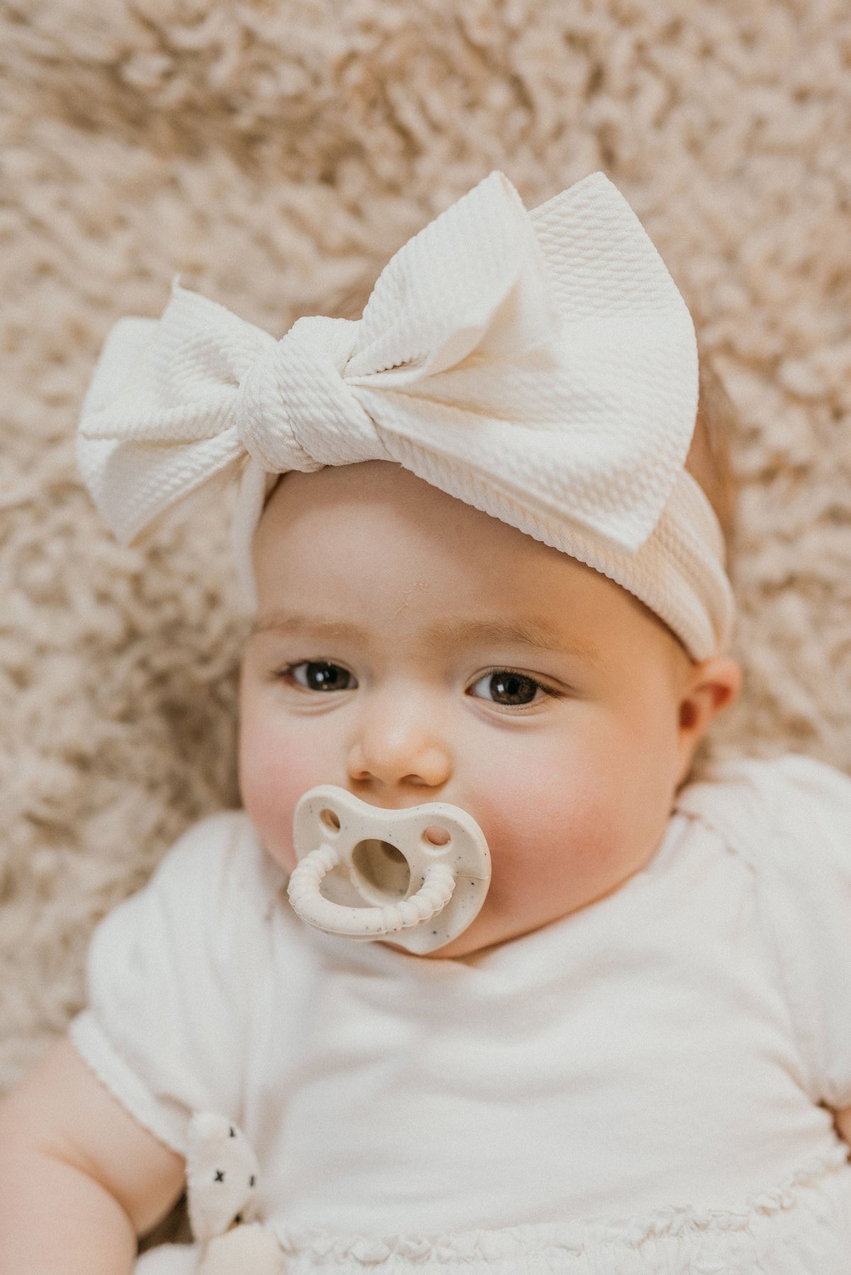 Baby wearing a white bow headband and pacifier on a soft beige background