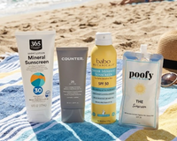 Four sunscreen products, including 365 Mineral Sunscreen, a Counter tube, Babo Botanicals spray, and a Poofy pouch, sitting together on a blue and yellow striped beach towel with the ocean in the background.