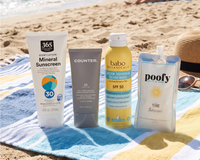 Four sunscreen products, including 365 Mineral Sunscreen, a Counter tube, Babo Botanicals spray, and a Poofy pouch, sitting together on a blue and yellow striped beach towel with the ocean in the background.