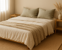 Minimalist bedroom with a neatly made bed, cream bedding, green pillows, a knitted throw, and natural wood furniture with soft natural light.