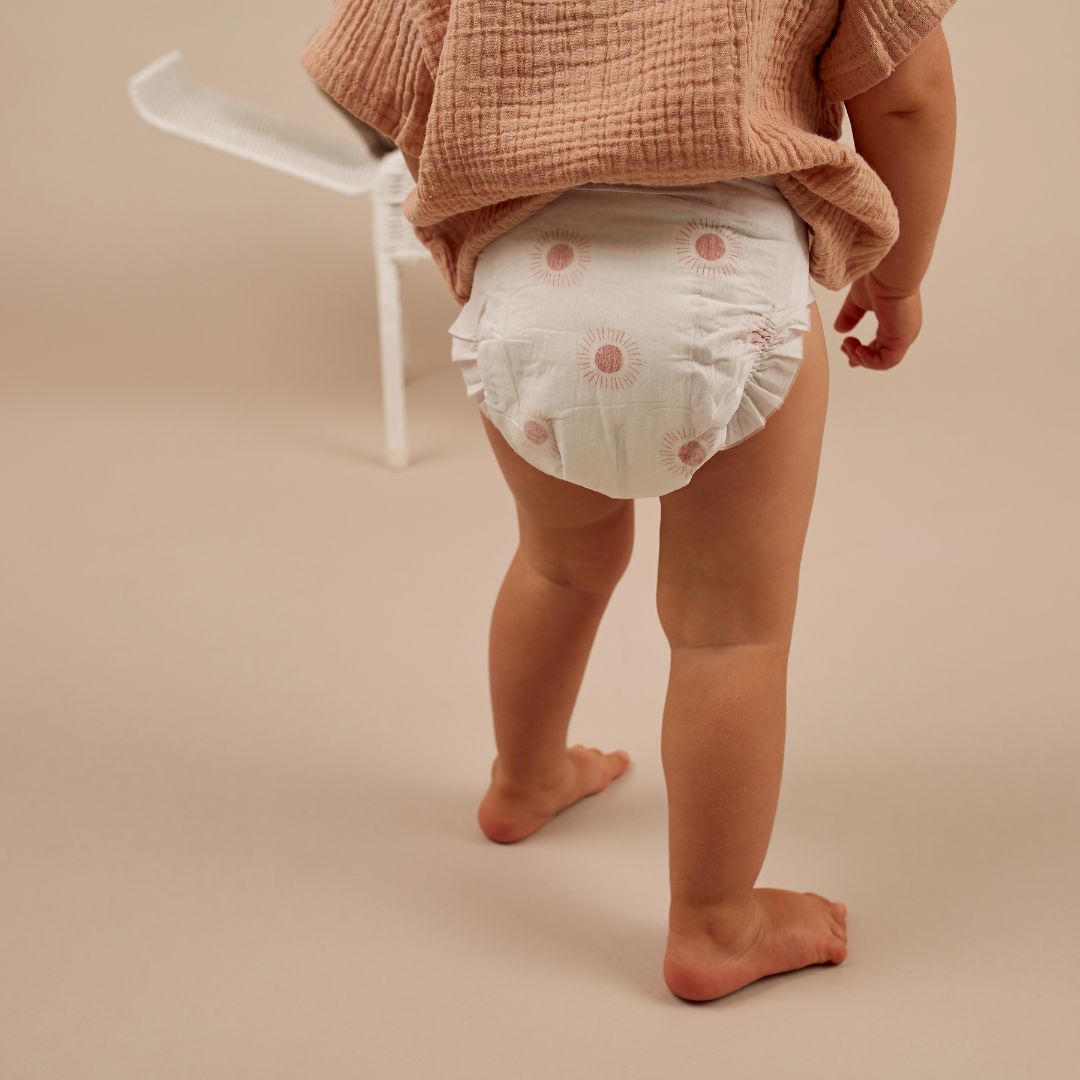 Toddler in a disposable diaper walking barefoot indoors, seen from behind.