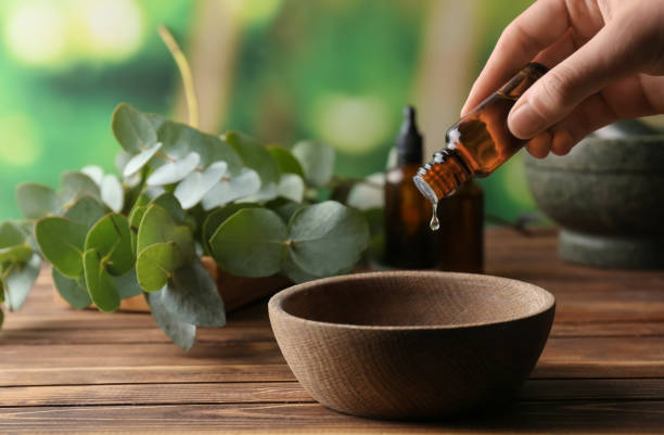 Hand pouring essential oil into a wooden bowl beside fresh eucalyptus leaves on a wooden table.
