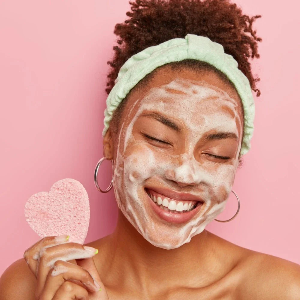 Smiling woman with a foamy facial cleanser on her face, holding a pink exfoliating sponge against a pink background.