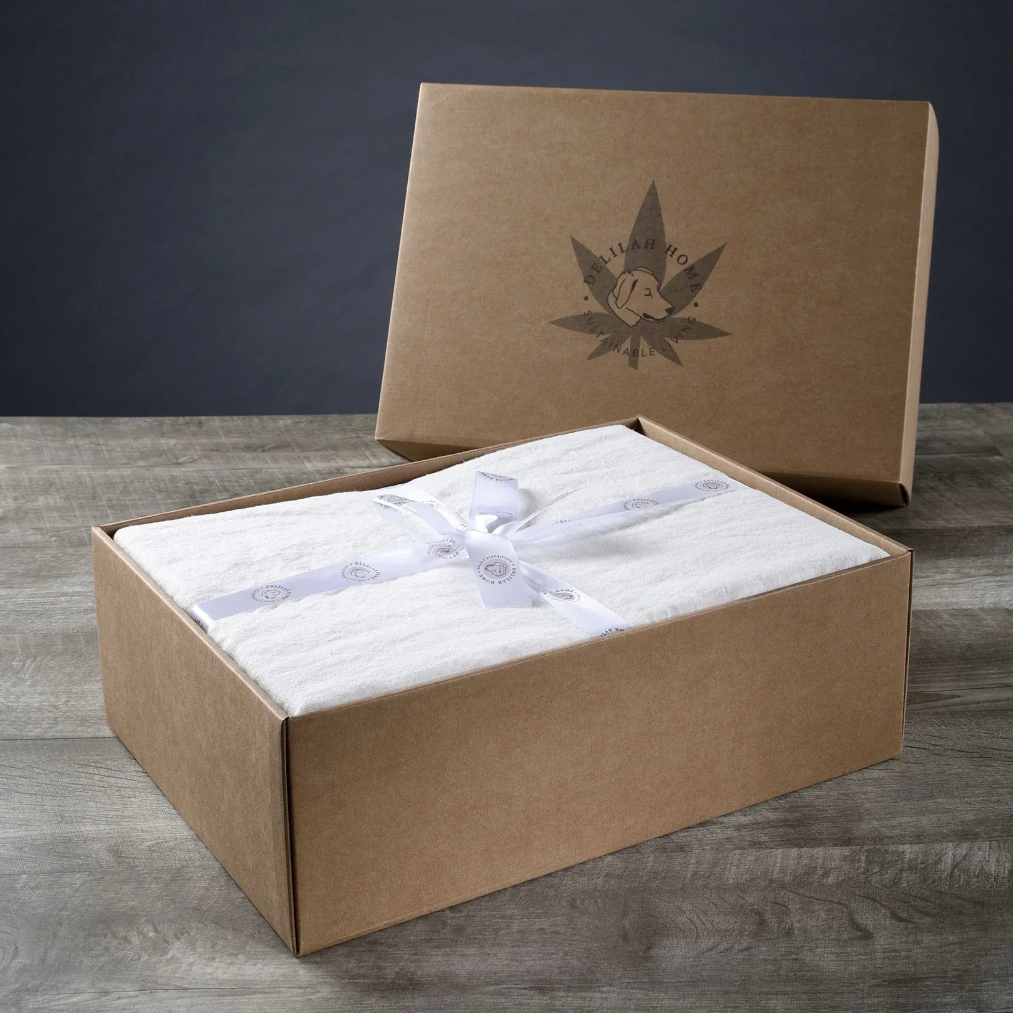 Open brown gift box with white fabric and ribbon on a wooden surface, featuring a cannabis leaf logo.