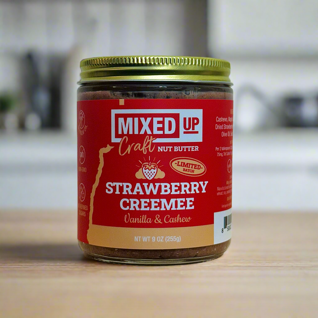 Jar of Mixed Up Craft Strawberry Creemee Nut Butter on a wooden surface with a blurred kitchen background.