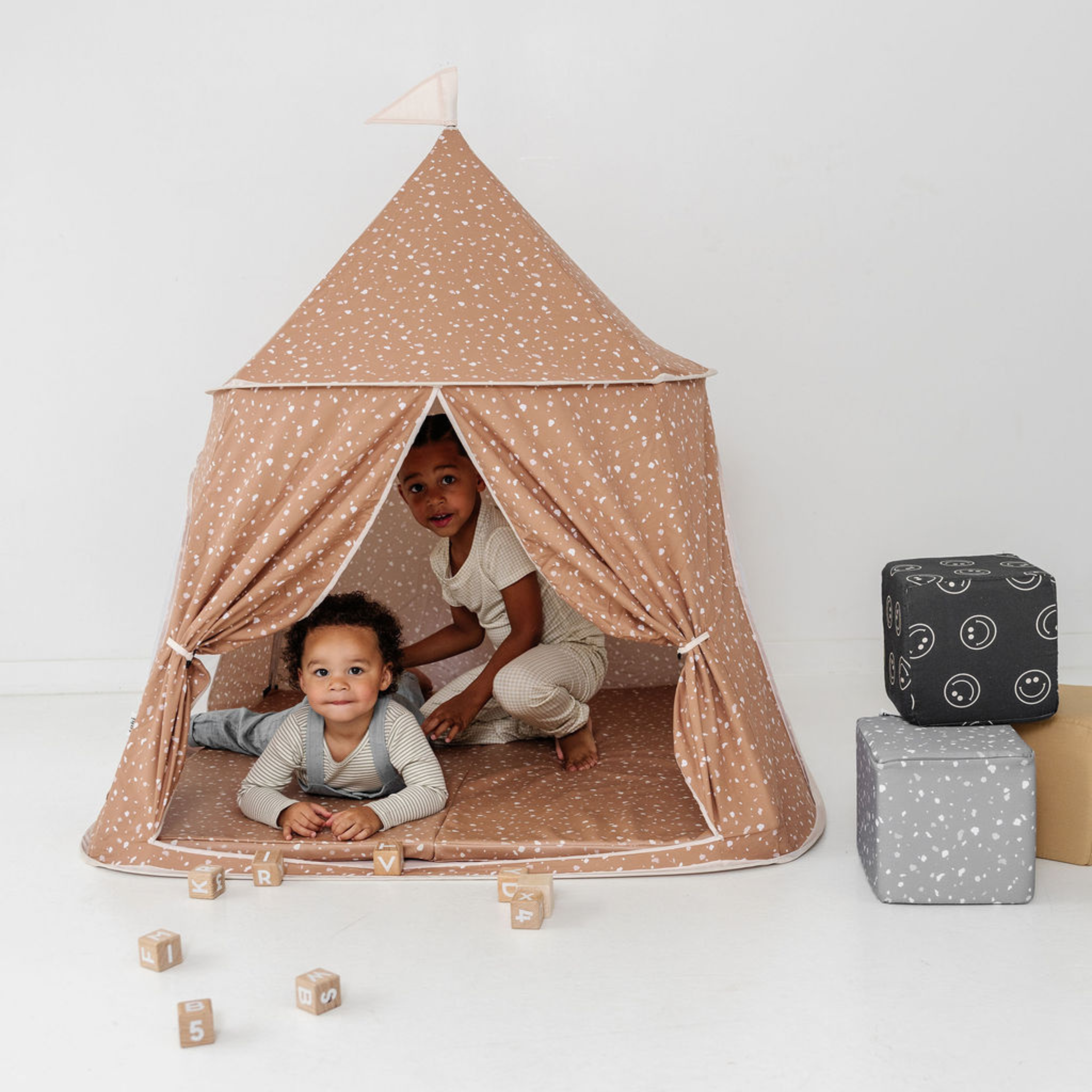 Two children playing inside a beige polka dot tent with blocks and boxes on a white background