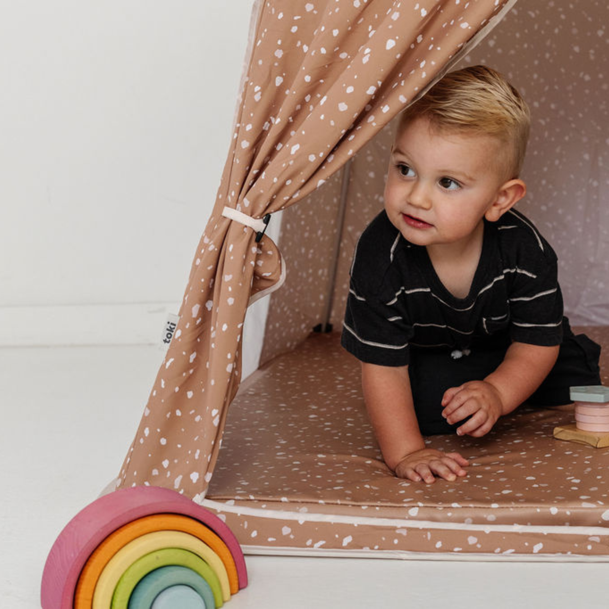 Child playing inside a homemade cardboard playhouse with rainbow toys.