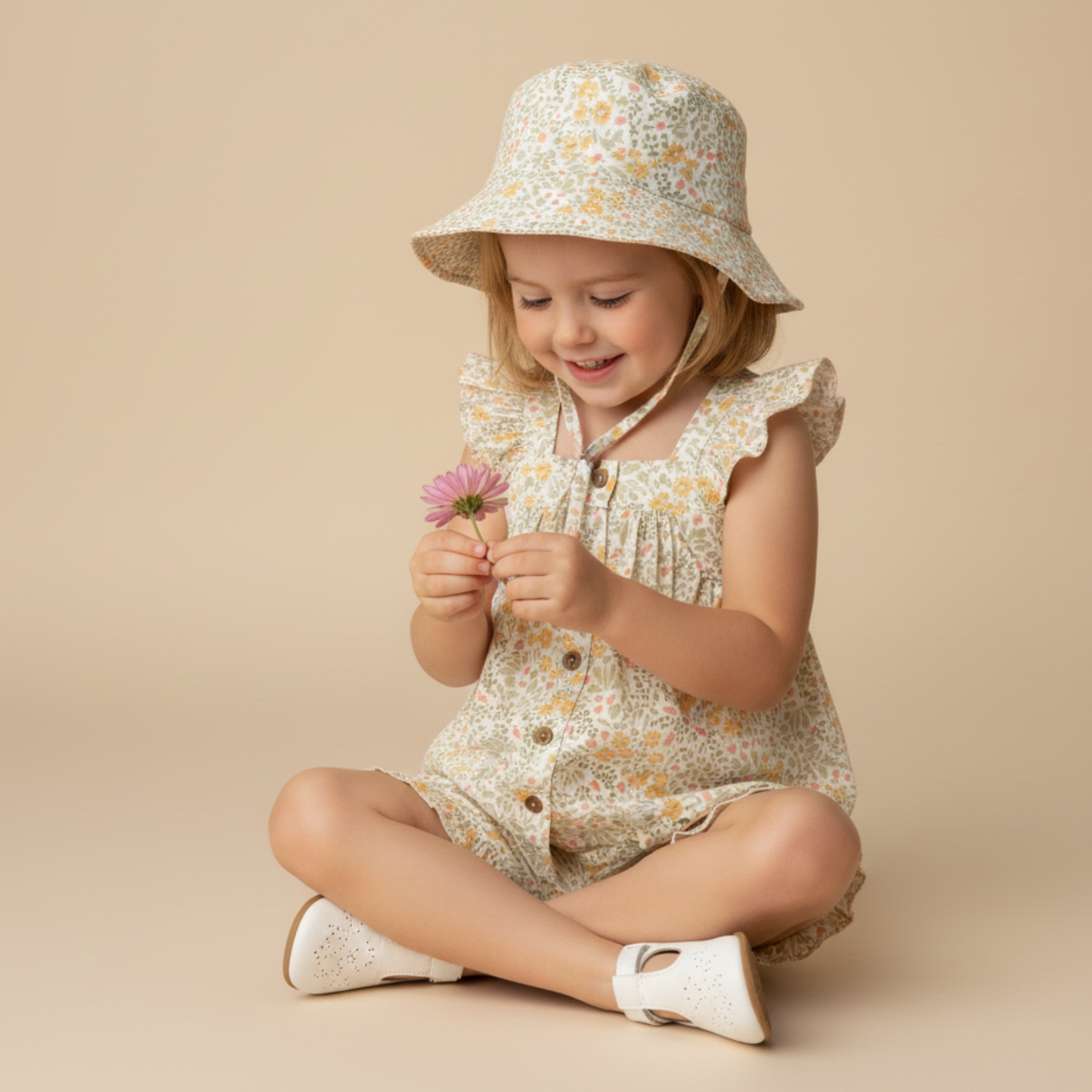 Child wearing a floral dress and hat, holding a flower on a beige background