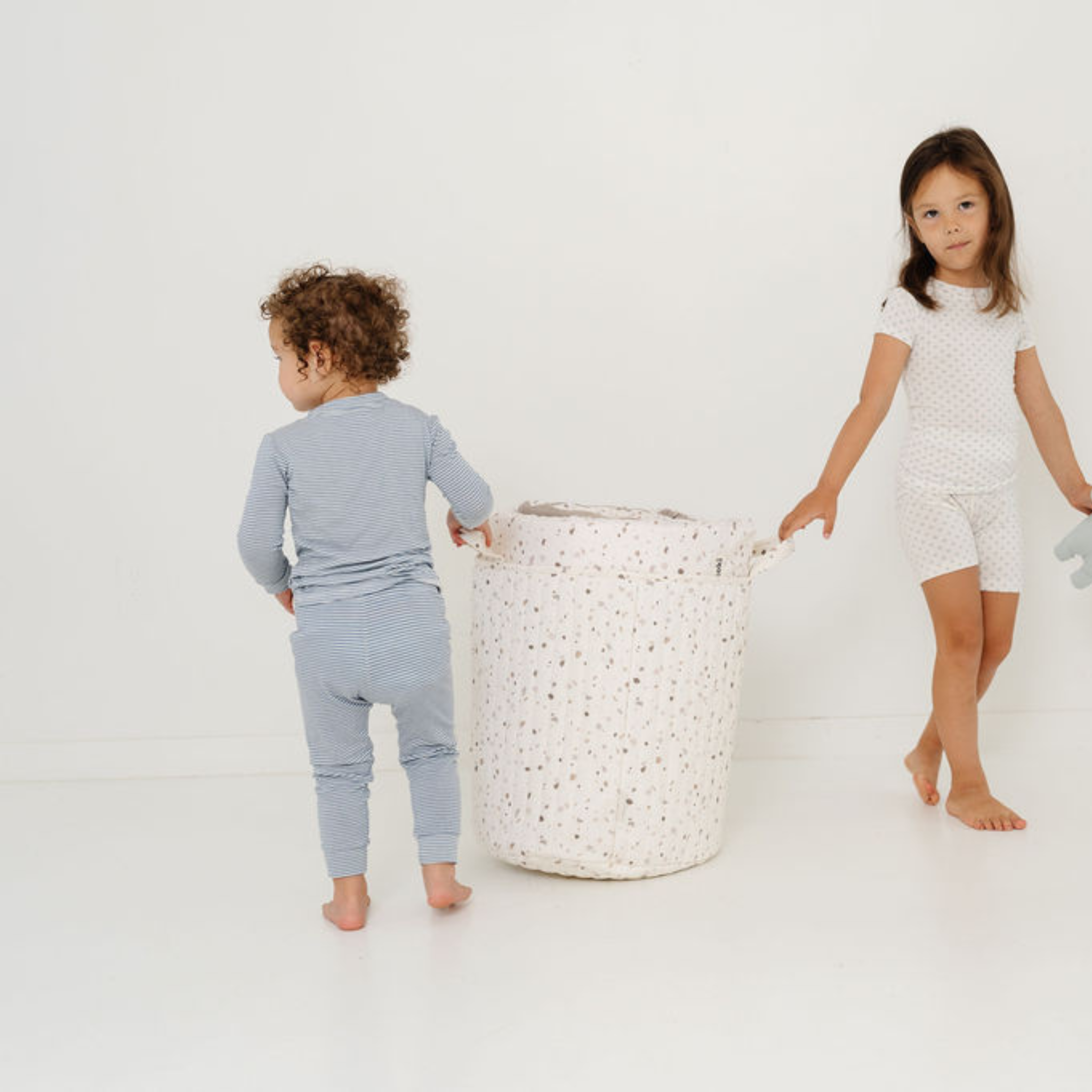 Two children in pajamas standing next to a laundry basket on a white background