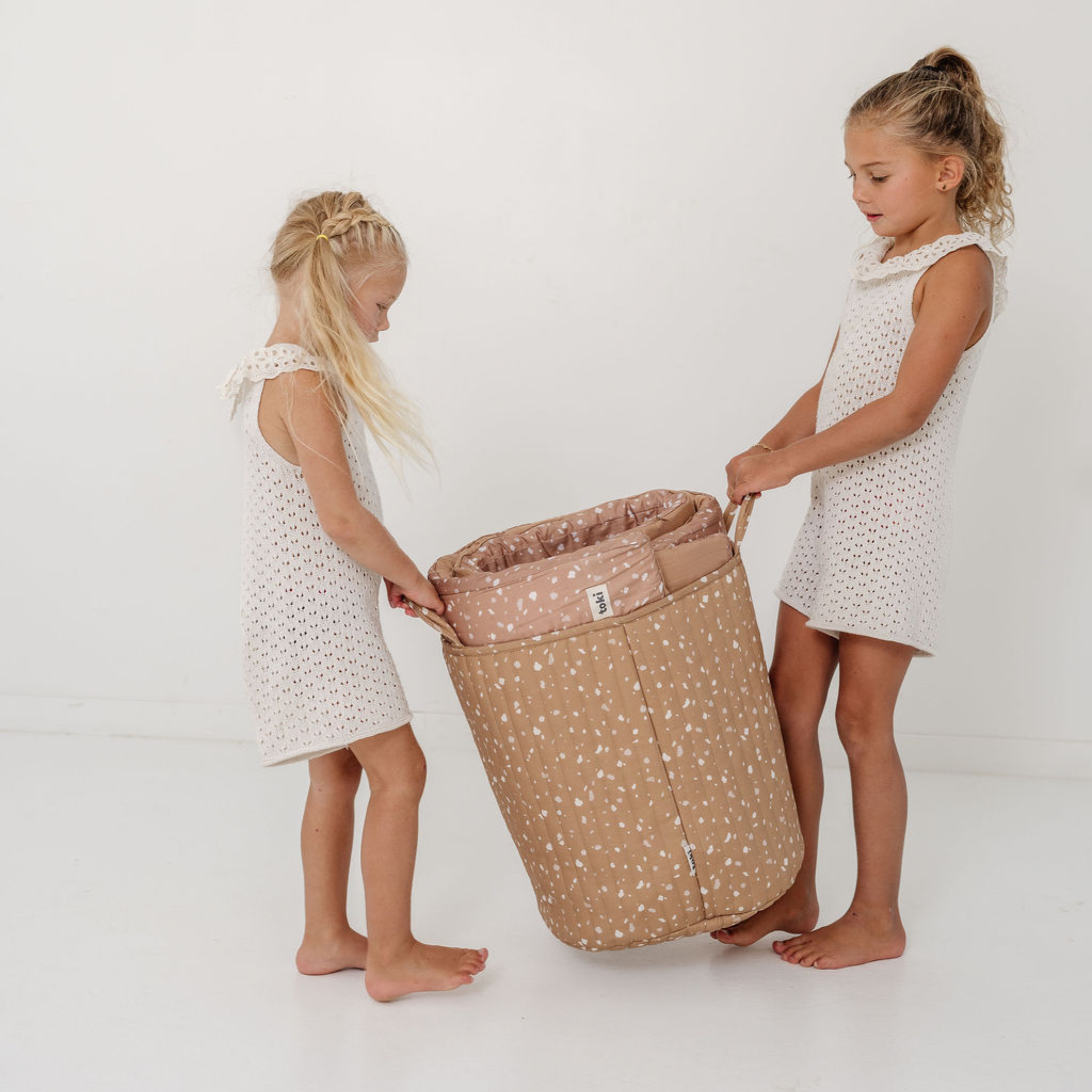 Two young girls holding a large beige laundry basket with a pink interior on a white background