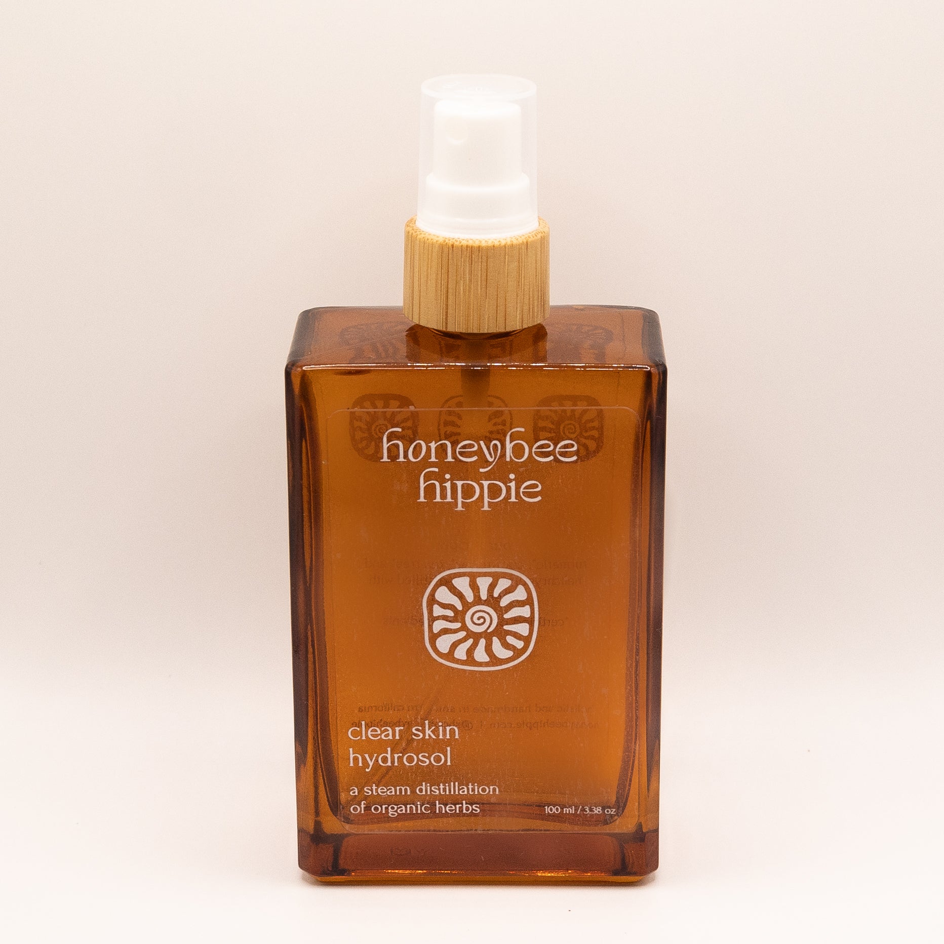 A rectangular amber glass bottle of 'honeybee hippie' Clear Skin Hydrasol steam distillation herbal spray with a bamboo-textured pump and white spray head, set against a plain light beige background.