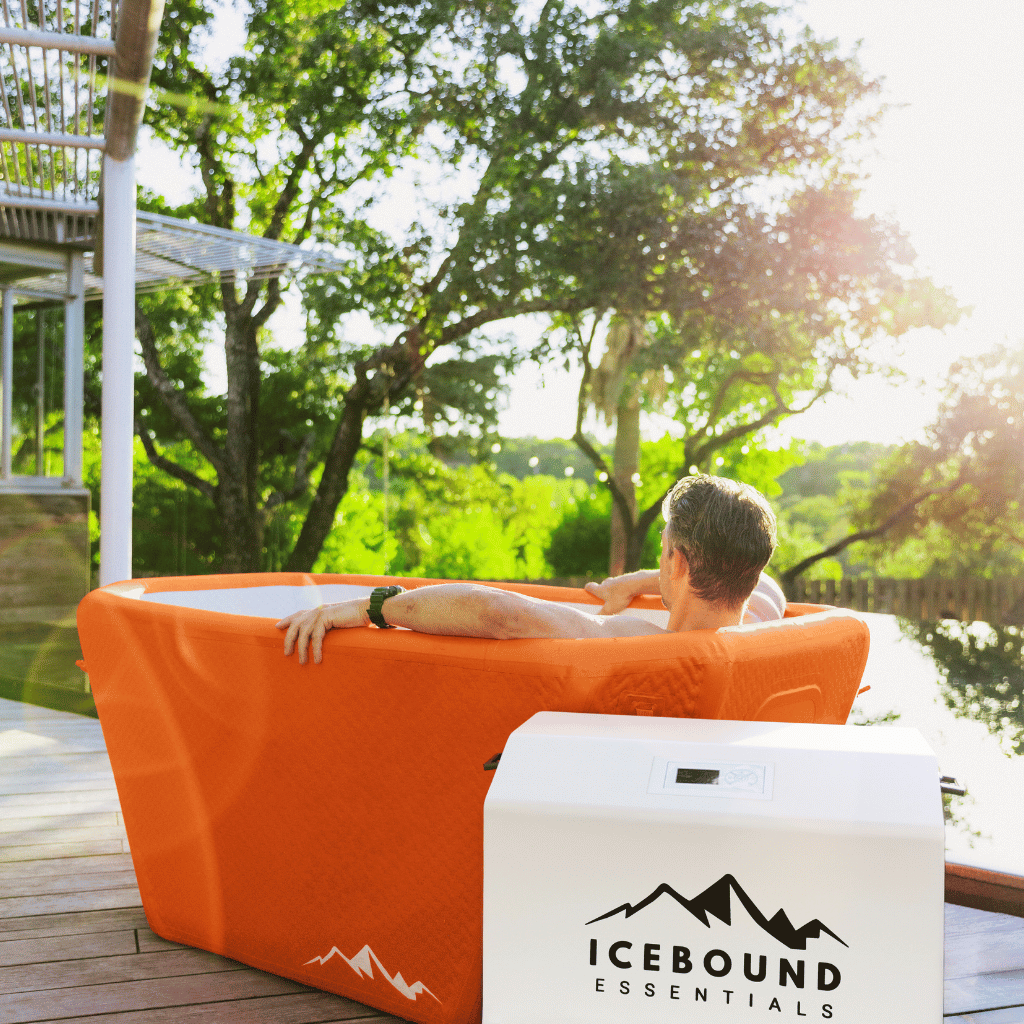 Person relaxing in an inflatable hot tub on a wooden deck with trees in the background, featuring Icebound Essentials branding.