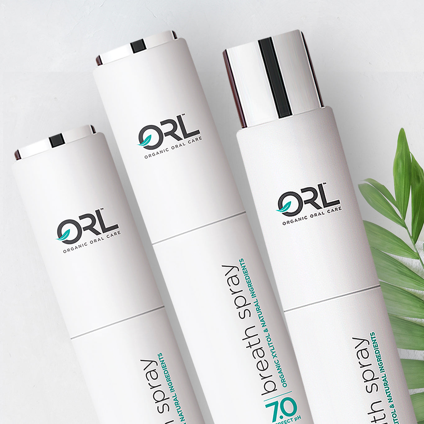 Three ORL Organic Oral Care breath spray bottles on a light background with green leaves.