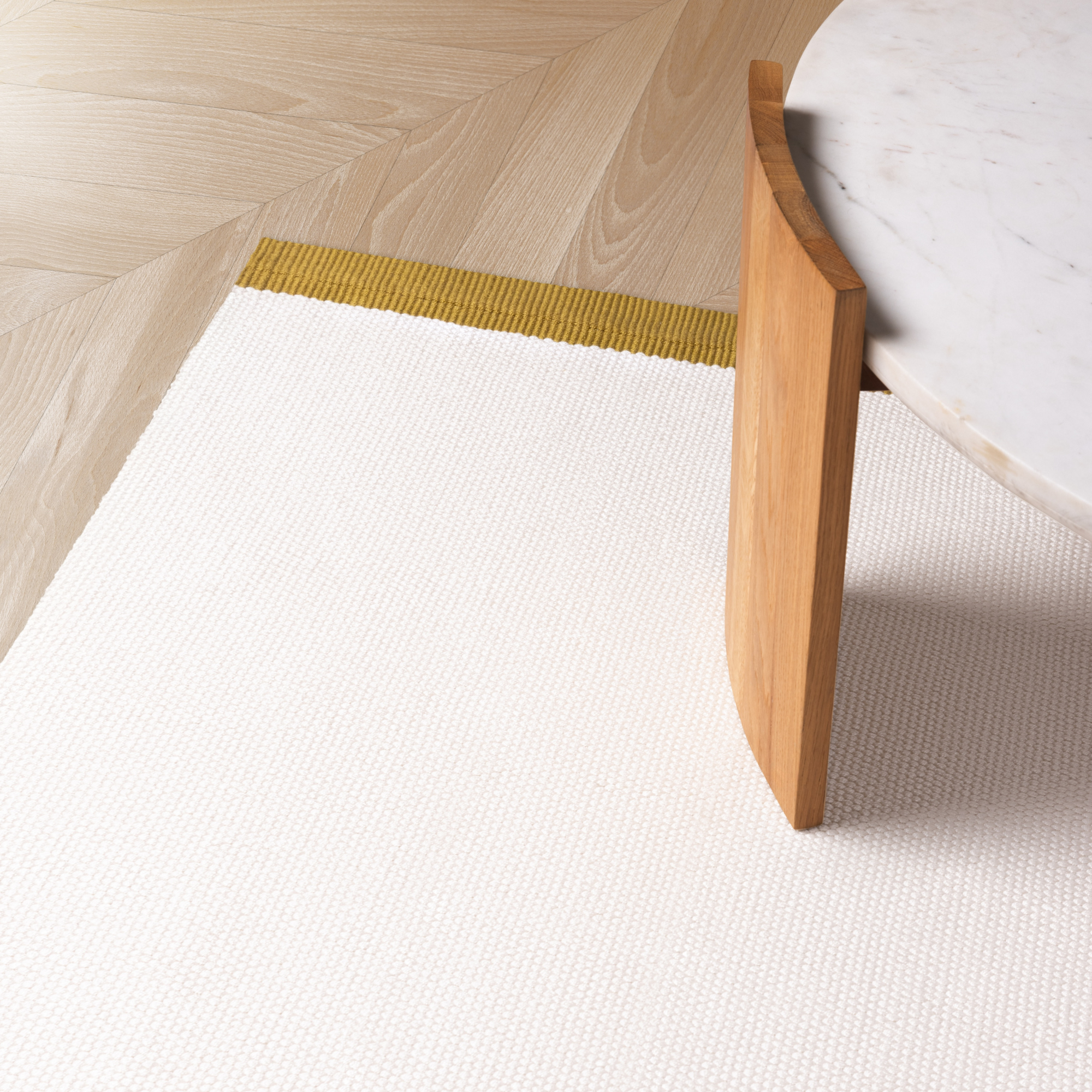 A close-up of a textured white rug with a mustard yellow stripe, shown in a modern room setting with a light wood herringbone floor and a marble-topped table leg.
