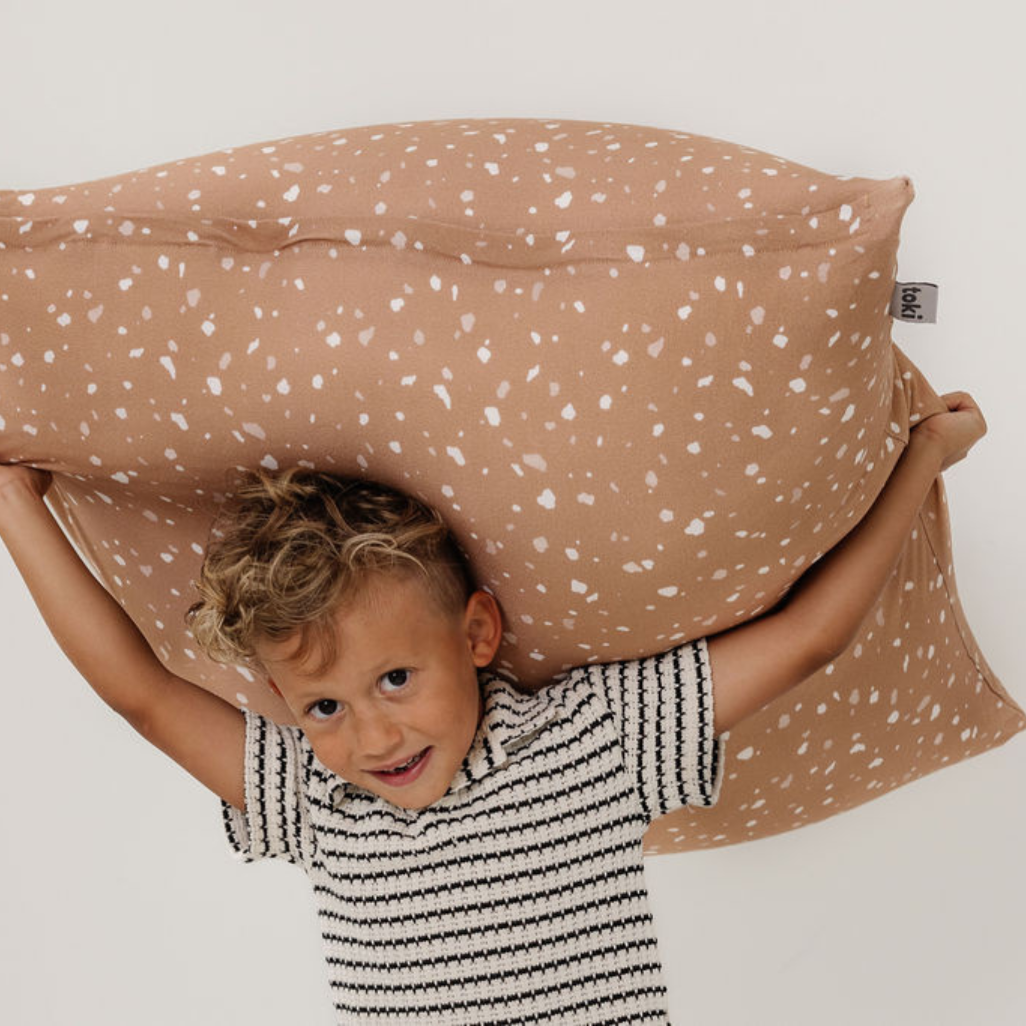 Child holding a large brown pillow with white speckles on a plain background