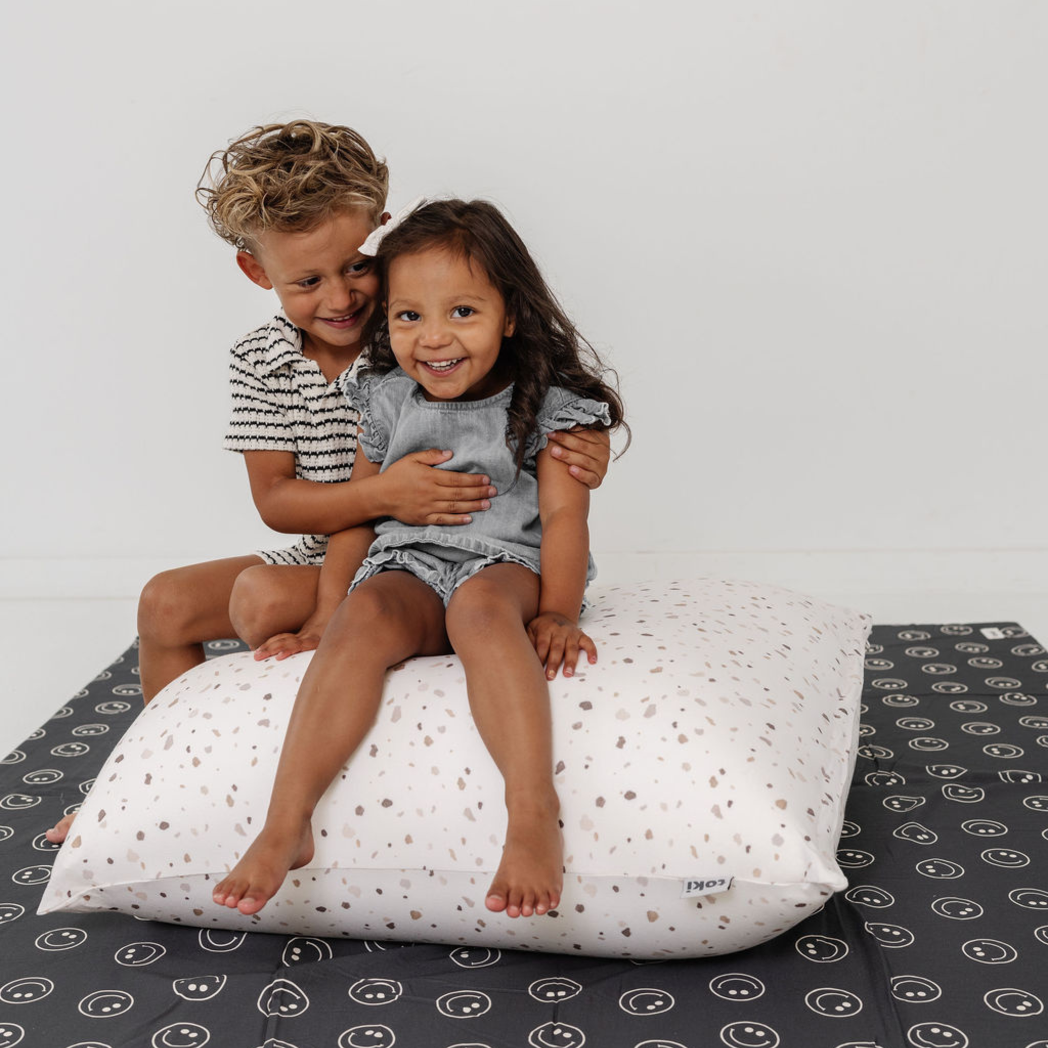 Two children sitting on a large pillow with a plain background