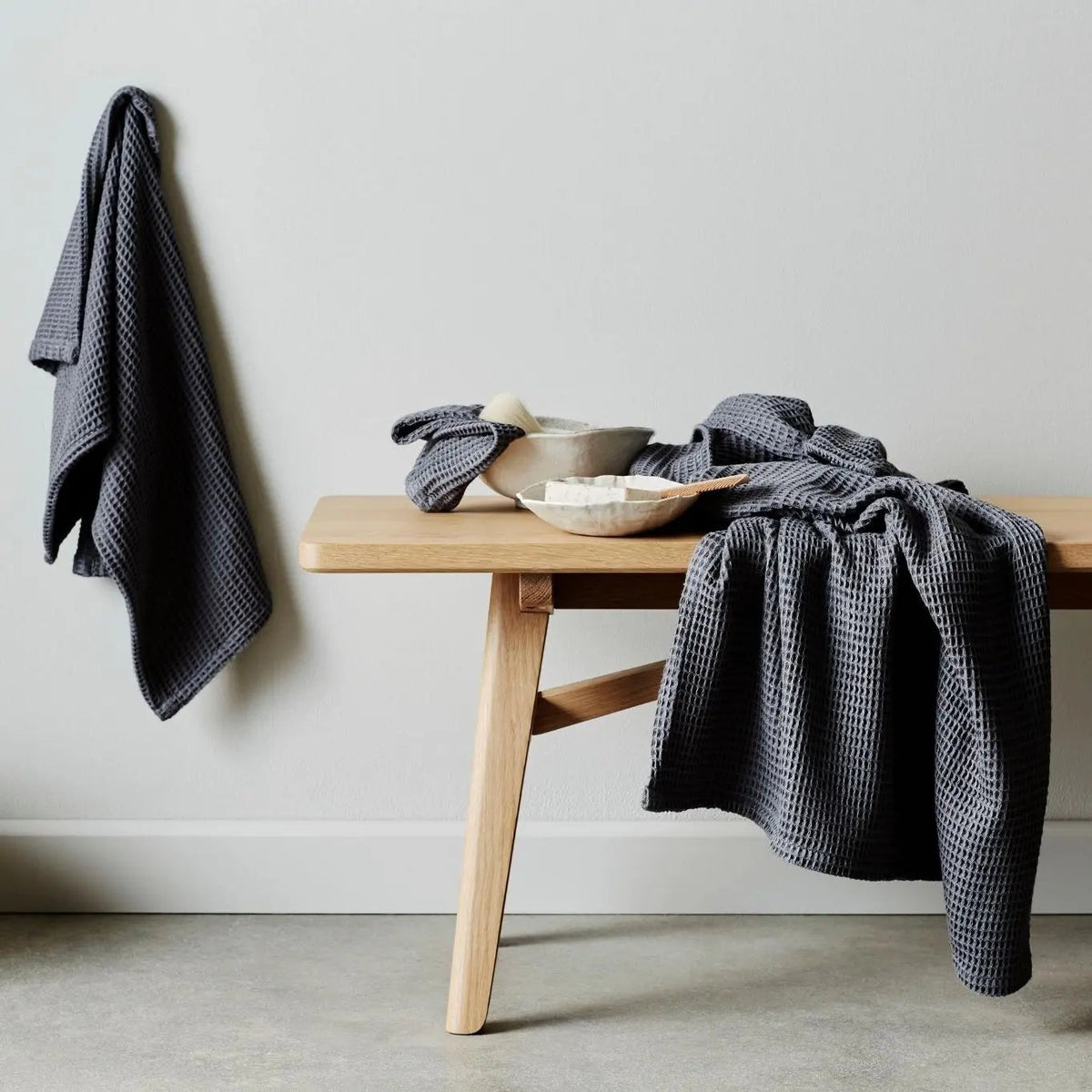 A dark gray waffle-weave kitchen towel draped over a light wood bench against a neutral gray wall.