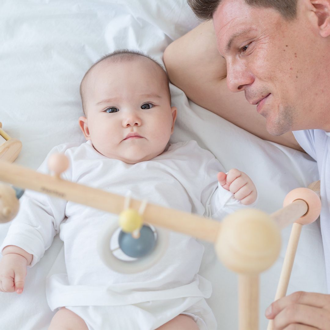 Baby lying on a white blanket with a wooden toy, next to a person holding the toy.