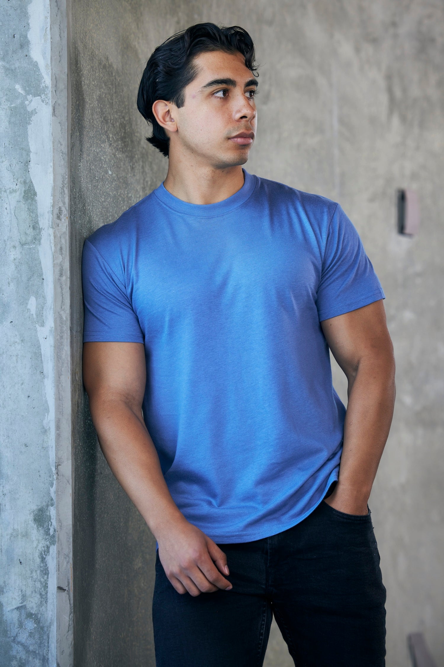 Man wearing a blue t-shirt leaning against a concrete wall.