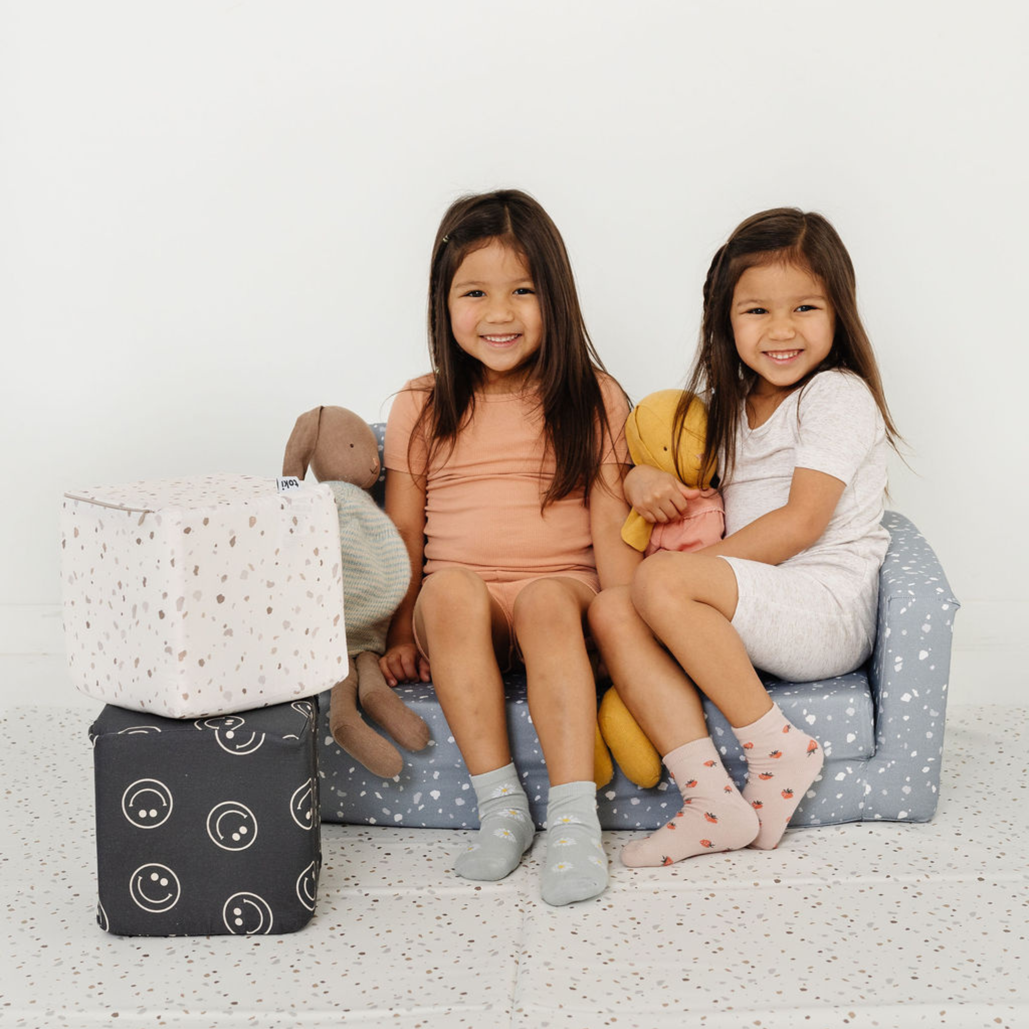 Two young girls sitting on a small blue chair with toys, surrounded by storage cubes.
