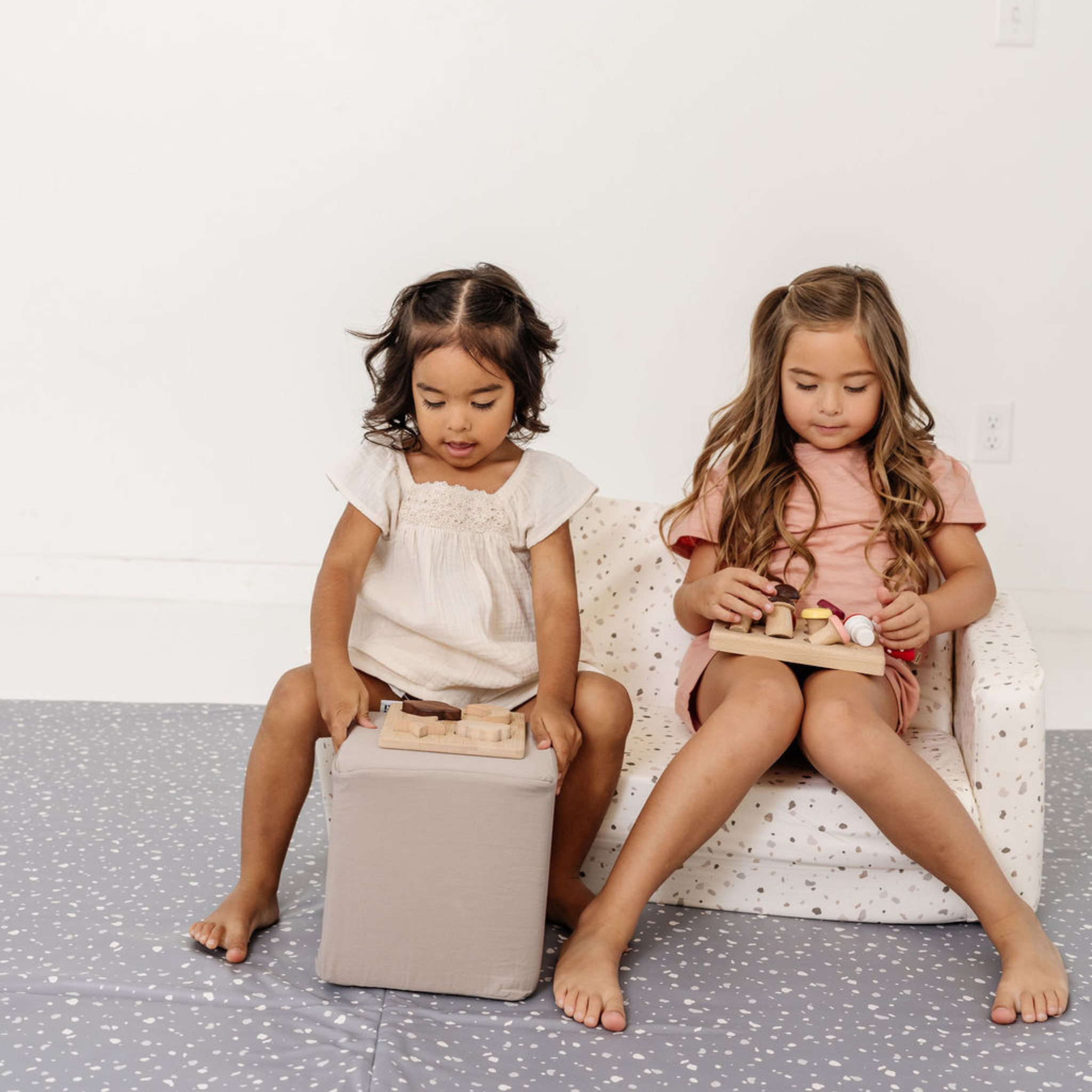 Two young girls playing with wooden toys on a white chair and gray floor.