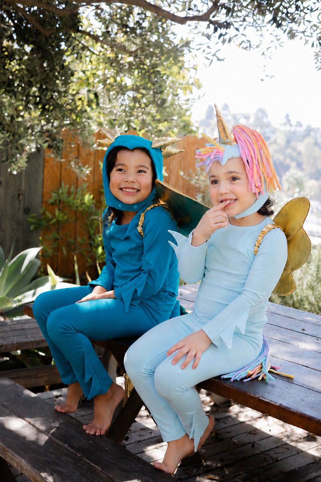 Two young children wearing matching unicorn-themed costumes sit on a wooden bench outdoors. The child on the left wears a teal hooded costume with gold horn details, and the child on the right wears a light blue long-sleeve costume with a pink, orange, and yellow horn and tail.