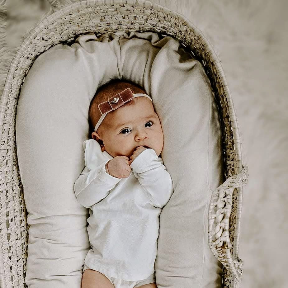 Baby in a Moses basket wearing a white outfit and headband with a bow.