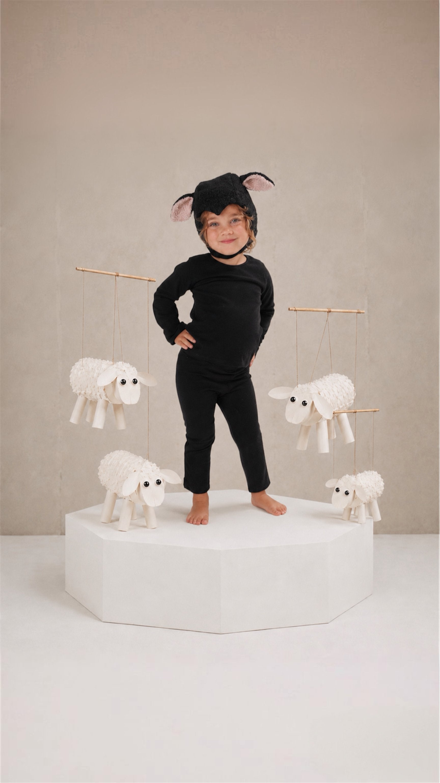 A child wearing a black long-sleeve outfit and a black mouse costume headband stands on a white pedestal, surrounded by hanging white sheep decorations against a neutral beige studio background.