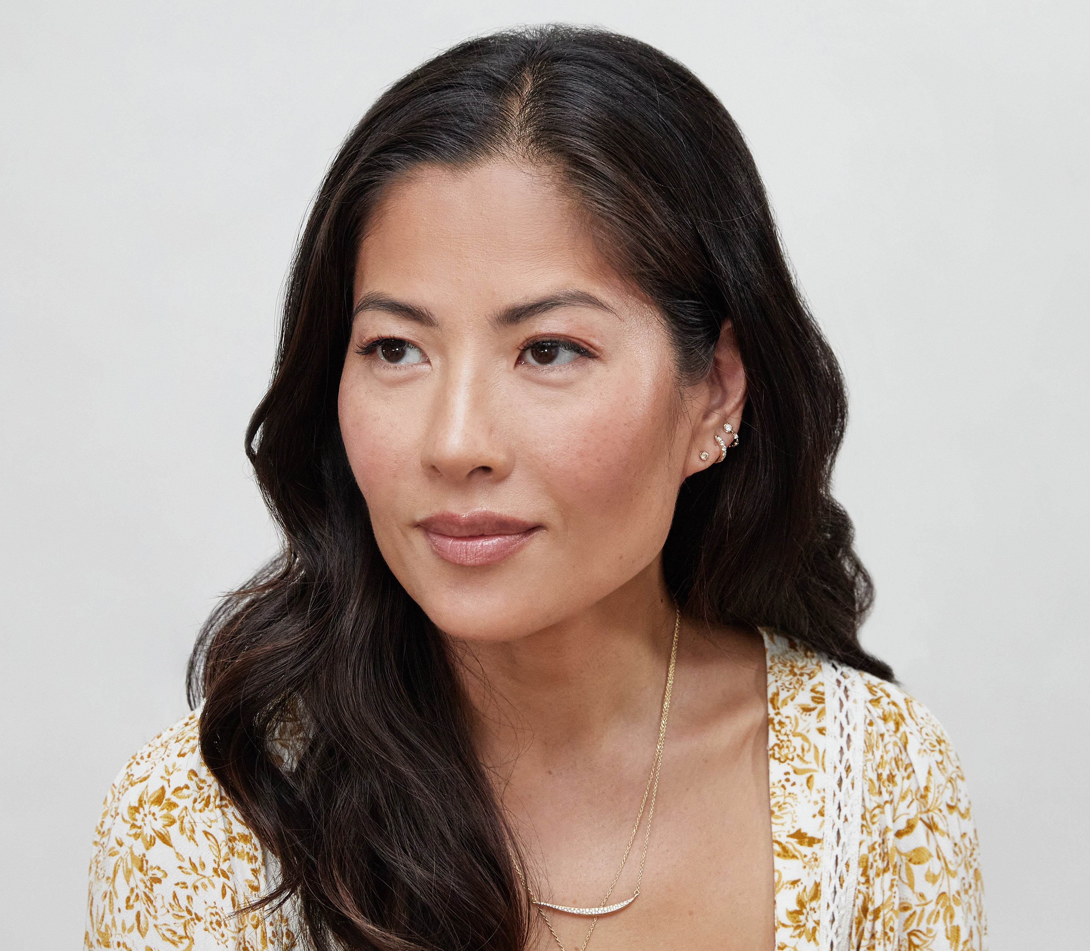 Woman with long dark hair wearing a gold necklace against a light background