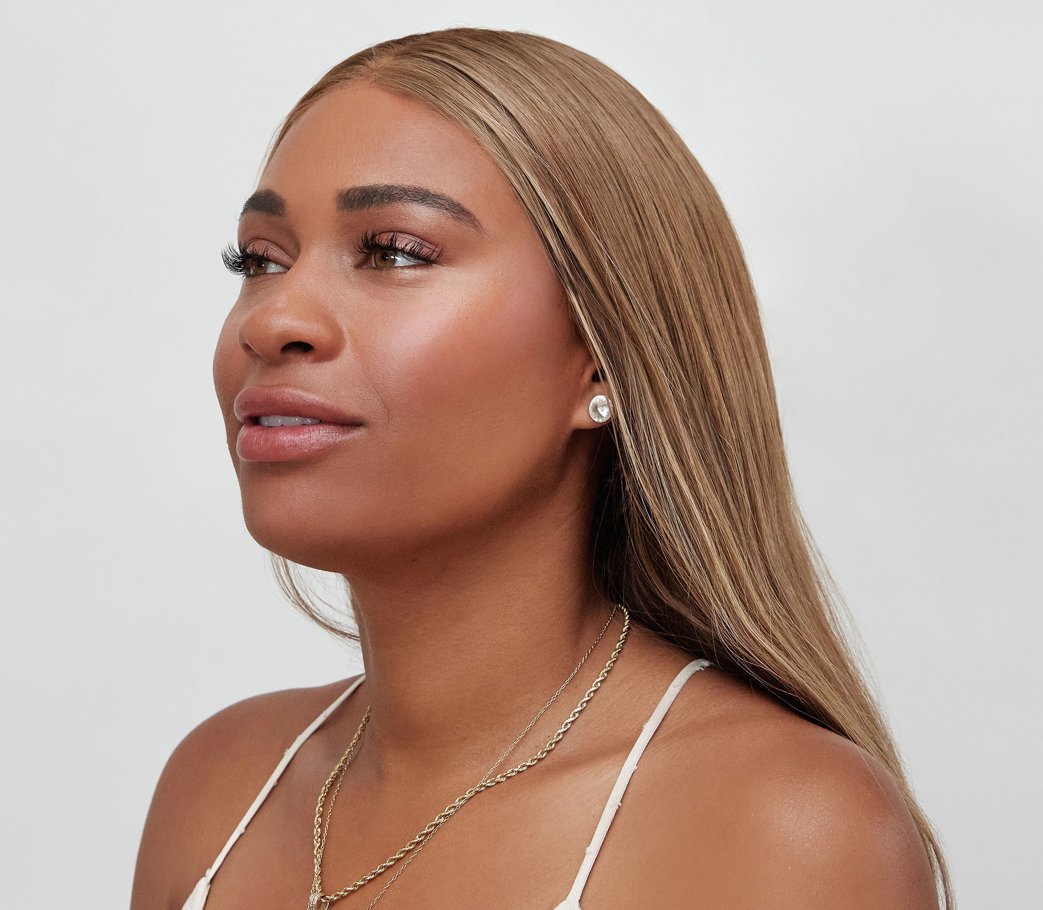 Woman with long blonde hair wearing a white top and gold necklace on a light gray background
