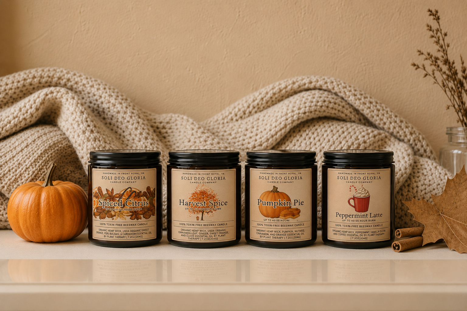 Four scented candles in dark glass jars with black lids are lined up on a white surface against a beige wall, with a knitted cream blanket behind them. A small orange pumpkin sits on the left, and dried leaves, cinnamon sticks, and a glass jar are placed around the candles.
