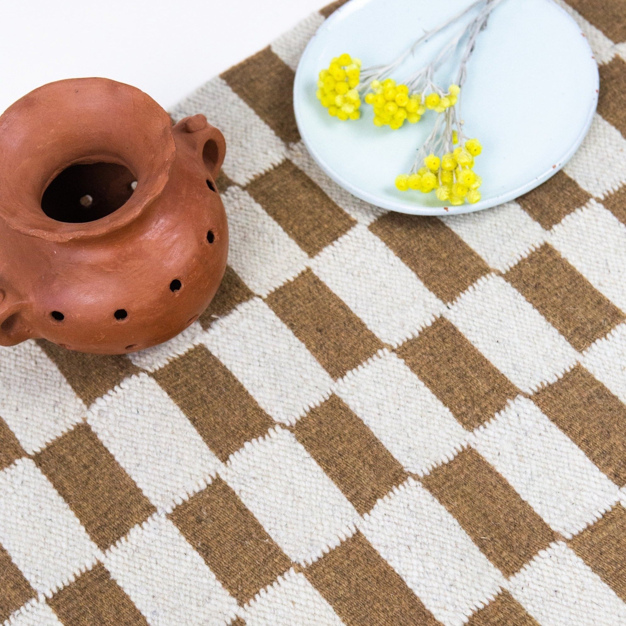 Checkered brown and white rug with a terracotta pot and white plate with yellow flowers on top.