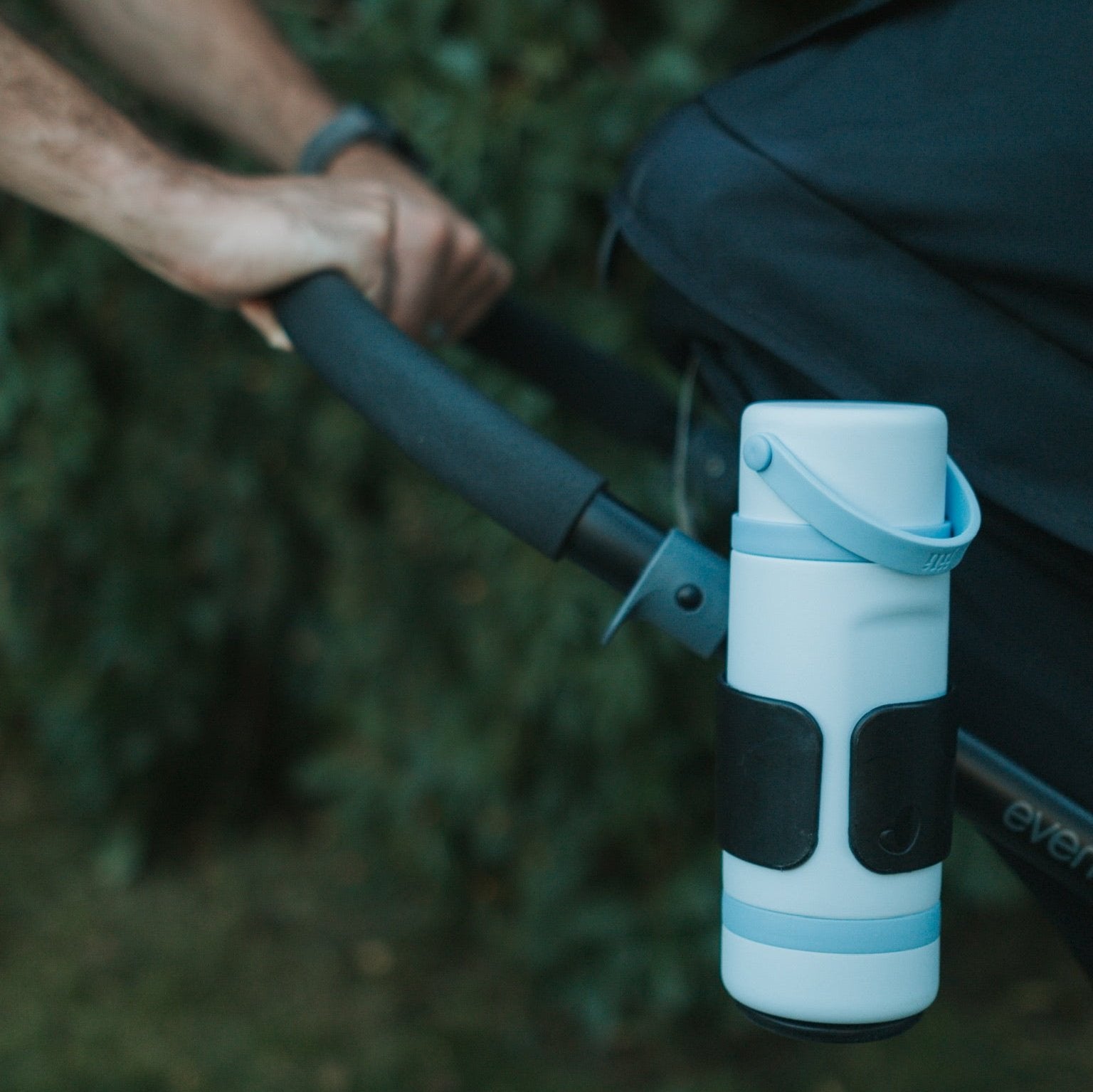 Light blue water bottle with black accents attached to a stroller handle, blurred green background