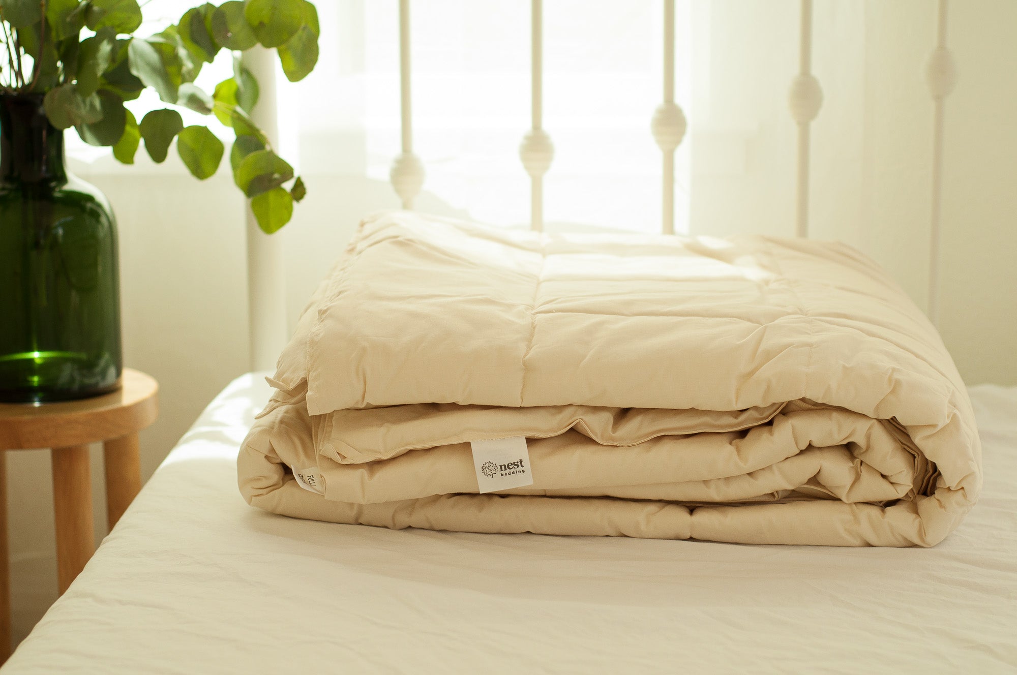 Beige comforter on a bed with a plant and side table in the background
