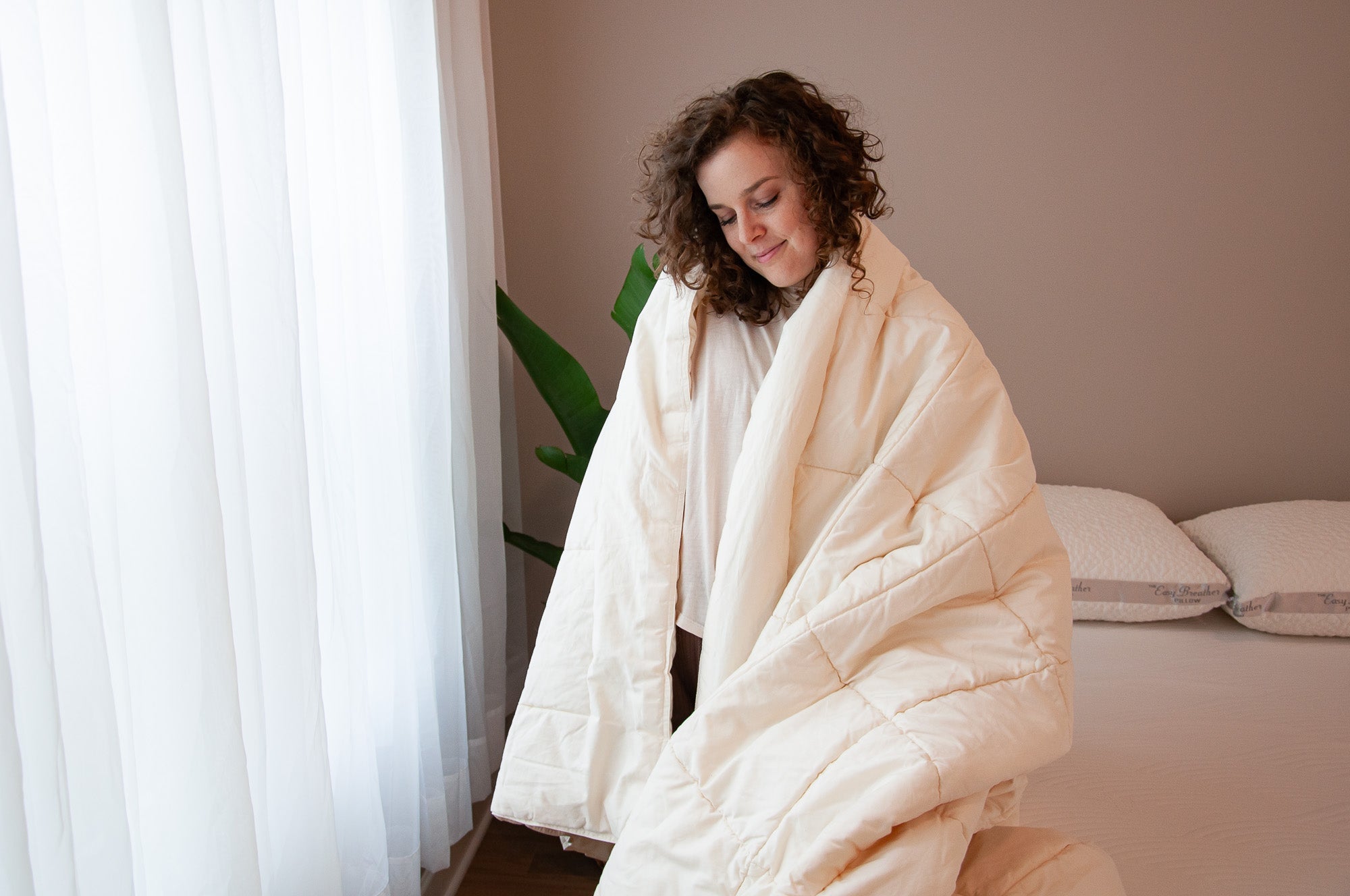 Woman wrapped in a white blanket sitting on a bed in a softly lit room.