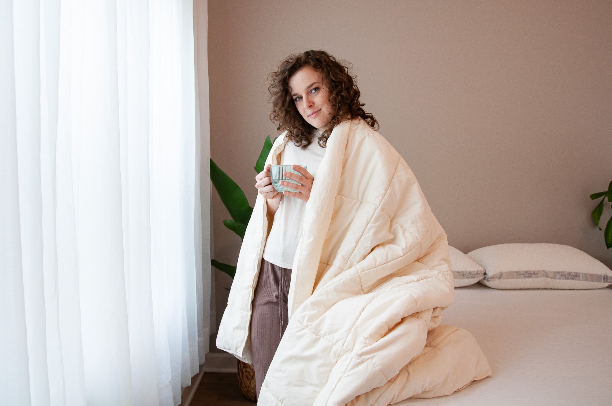 Woman wrapped in a beige blanket holding a mug in a cozy room.