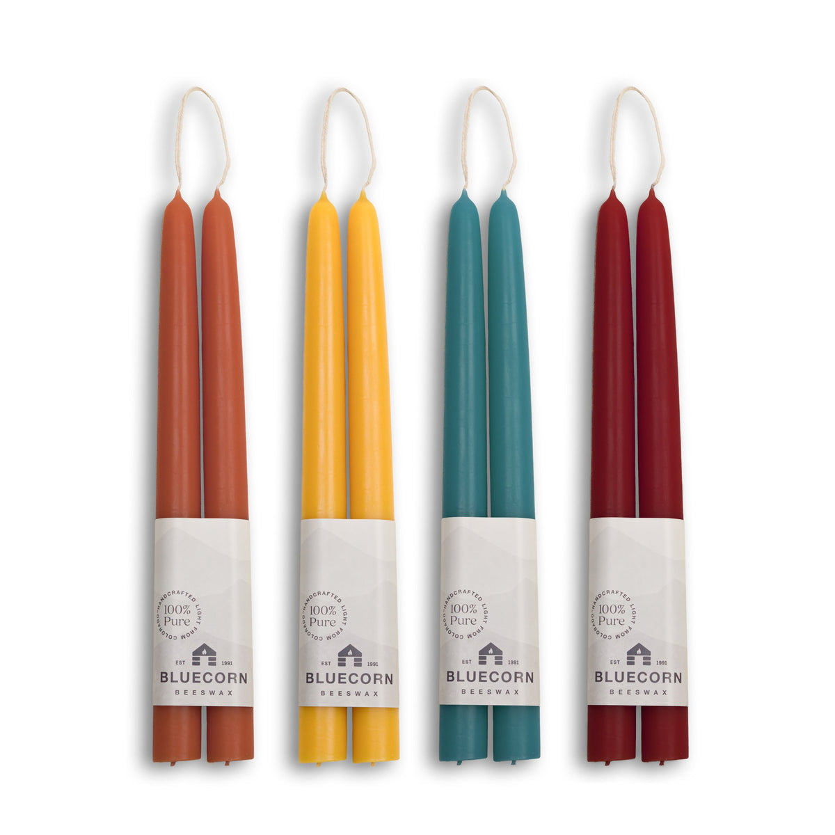 Four double taper candles in assorted colors (rust orange, mustard yellow, teal, and burgundy) with white paper bands labeled 'BLUECORN Candles' and white wicks, arranged vertically on a white background.
