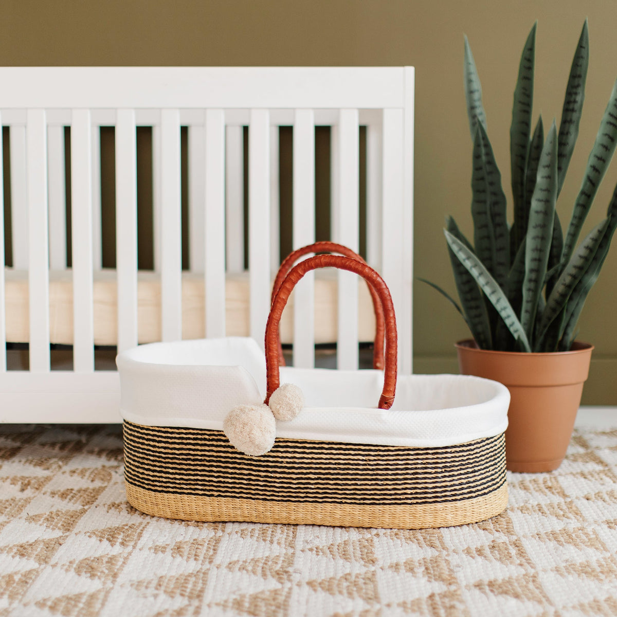 Woven basket with a red handle in front of a white crib with a plant in the background.