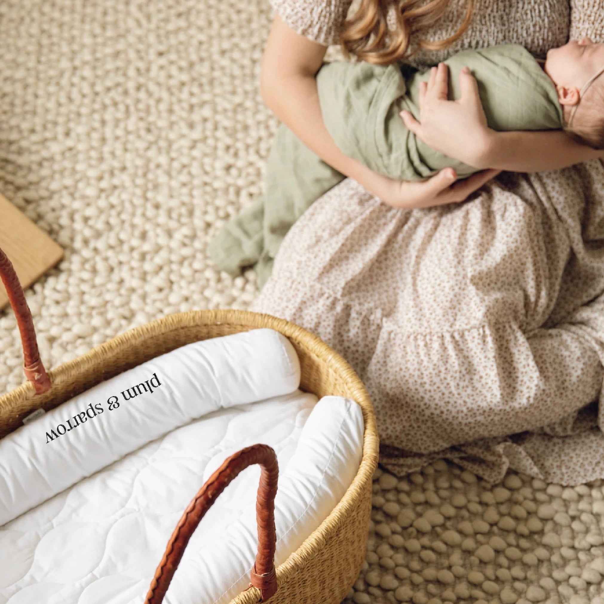 Person holding a baby wrapped in a green blanket next to a woven basket with white bedding.