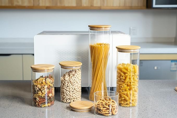 Glass storage jars with wooden lids on a kitchen counter, containing various types of pasta and grains.