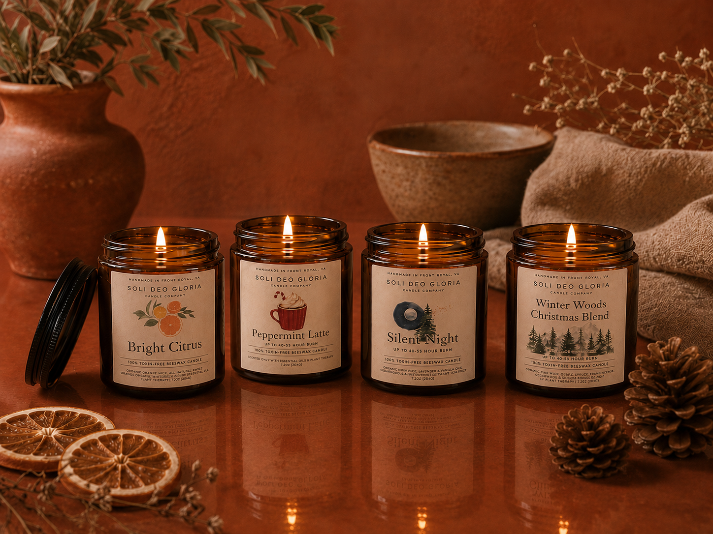 Four lit amber glass scented candles from Soul of the Savannah are arranged on a reflective surface with dried orange slices, a bowl, fabric, and pinecones in a warm rustic setting.