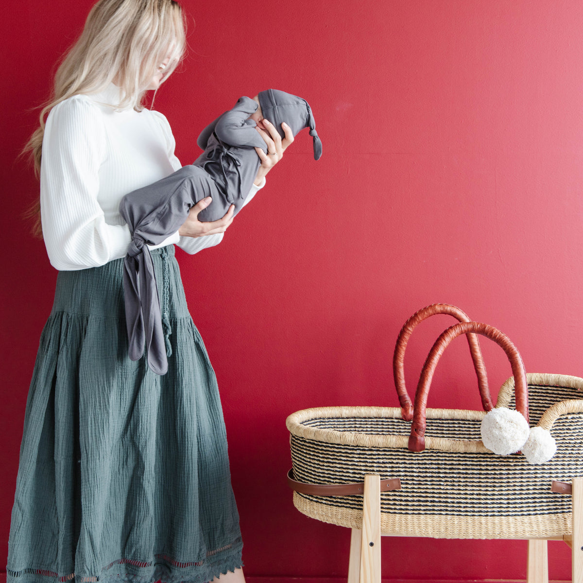 Woman holding a gray baby outfit against a red wall with a crib and basket in the foreground.
