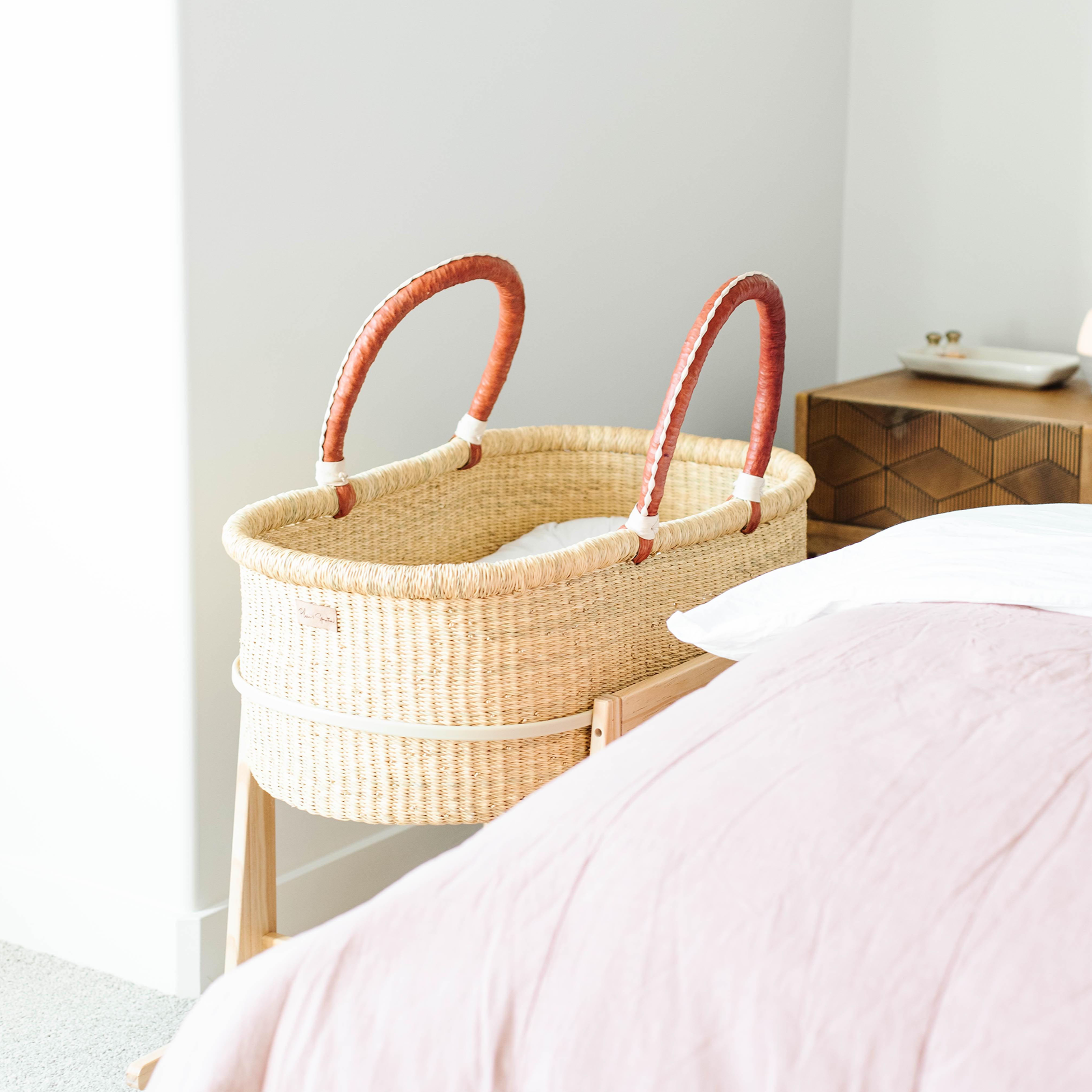 Woven baby bassinet with red handles next to a bed in a bedroom setting.