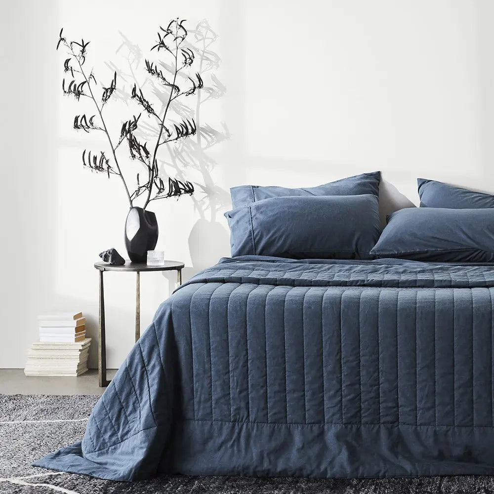 Blue quilted cover and pillows on a bed in a bedroom setting with a side table and decorative plant.